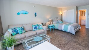 Una sala de estar con un sofá color crema, cojines verde azulado y azul, una mesa de centro de vidrio, y una cama con ropa de cama a rayas.
