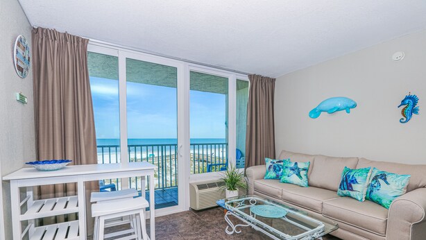 Un balcón con vistas al mar, una mesa blanca con sillas, un sofá color beige con almohadas azules y una mesa de centro de vidrio.