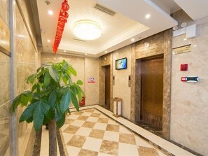 Un hall d’hôtel avec un sol à carreaux, une grande plante en pot, un ascenseur et une décoration rouge suspendue.