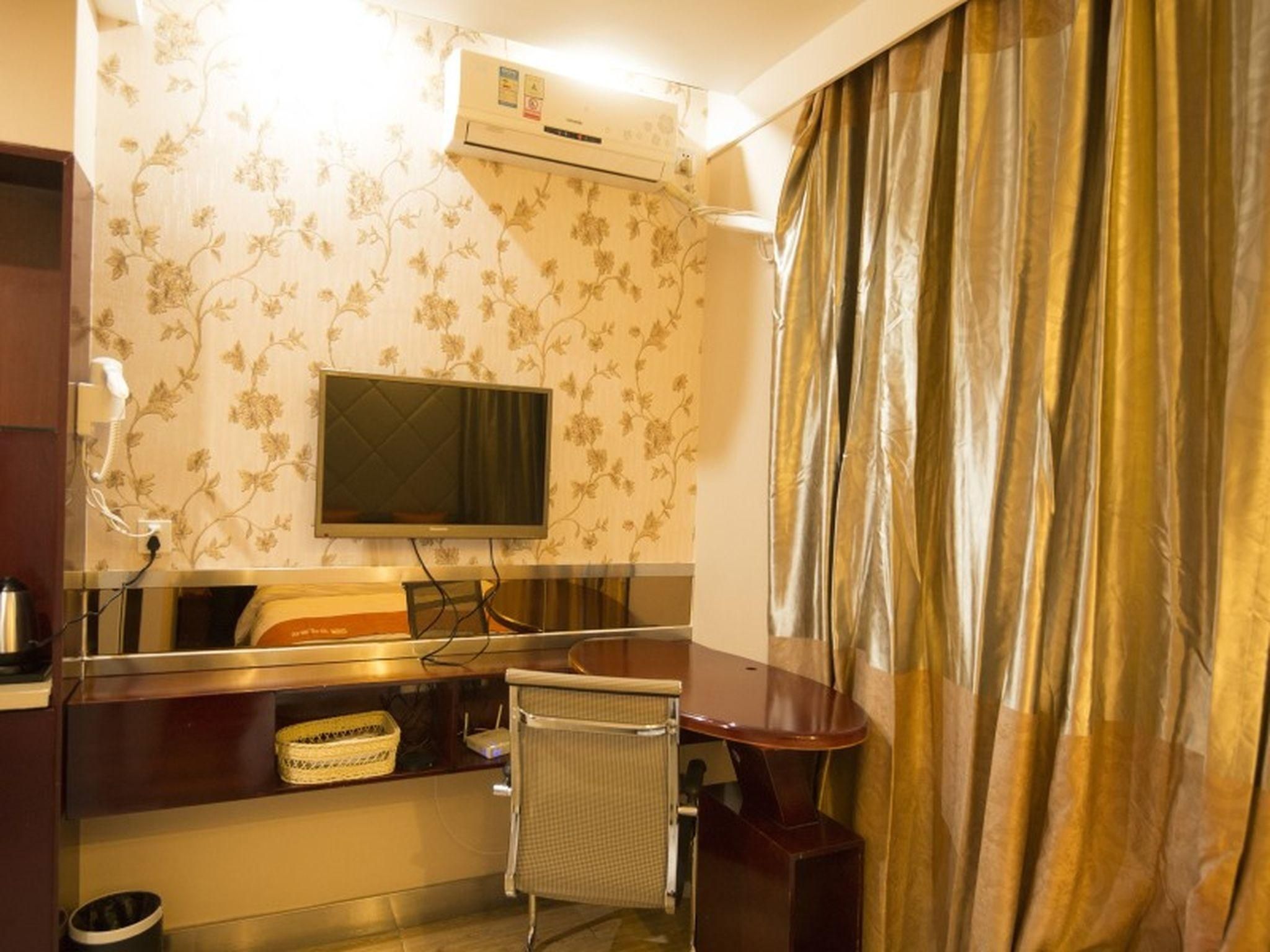 A hotel room with a flat-screen TV mounted on the wall, a desk with a chair, a small table, and a curtain.
