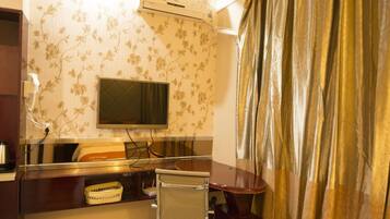 A hotel room with a flat-screen TV mounted on the wall, a desk with a chair, a small table, and a curtain.