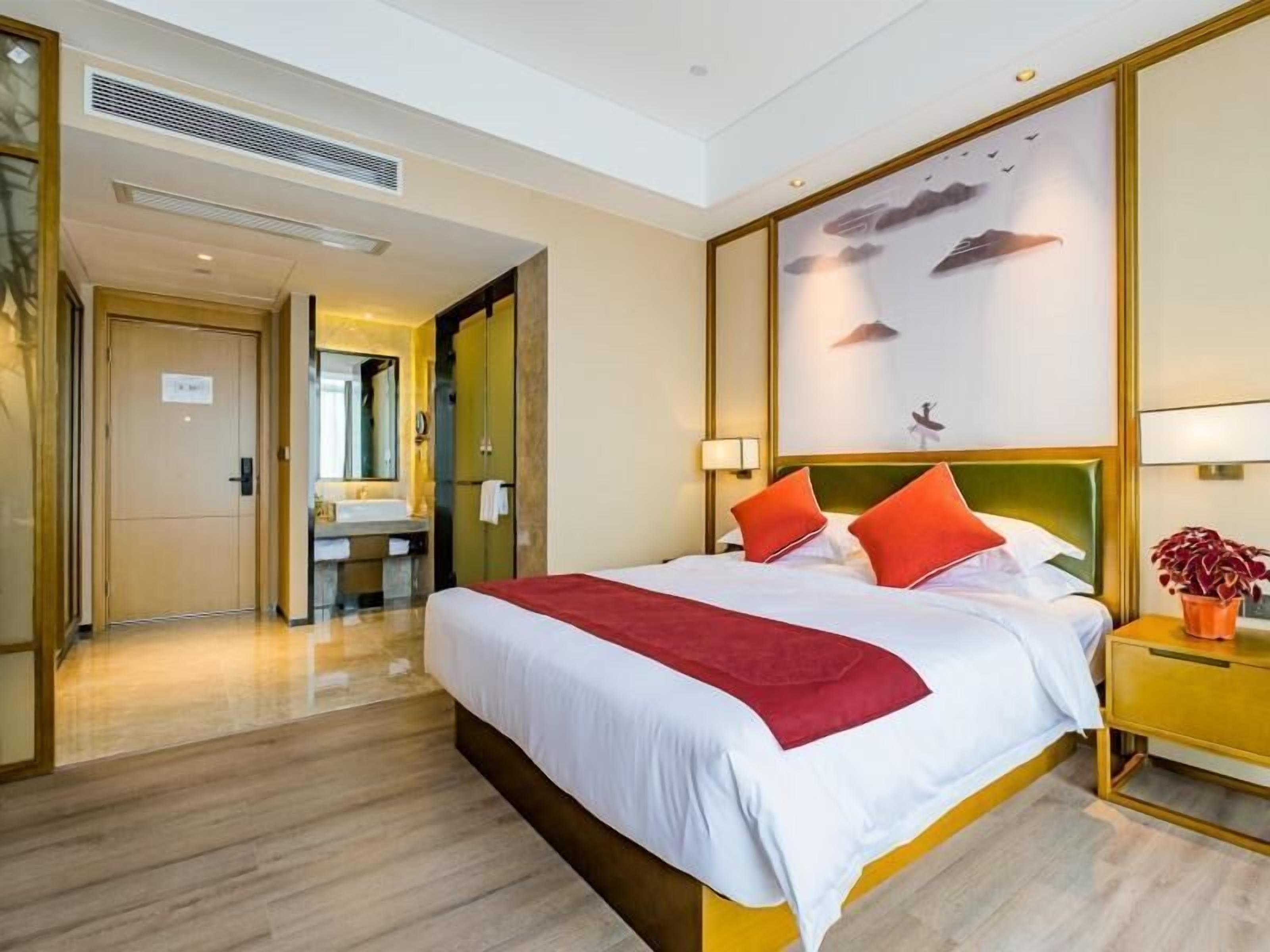 Une chambre d’hôtel avec un grand lit, une table de chevet avec une lampe, une œuvre d’art accrochée au mur et une salle de bain visible à travers une porte ouverte.