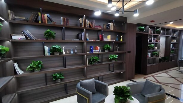 A modern library with dark wood bookshelves, multiple levels, and green plants. There are two grey armchairs and a small round table with a plant.