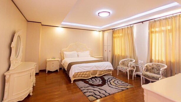 Habitación de hotel con cama, mesitas de noche, cómoda, espejo, silla, una mesita pequeña y cortinas.