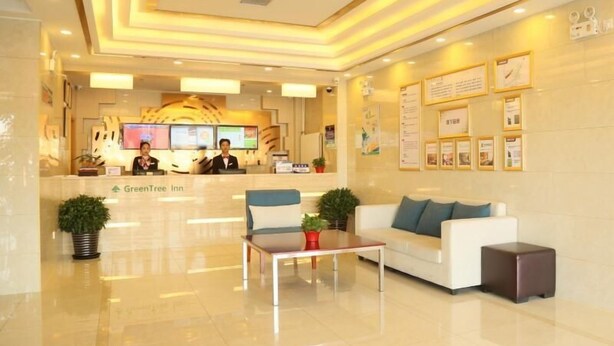 Un hall de hotel con una mesa de recepción, dos recepcionistas, una mesa de centro, un sofá y plantas en macetas.