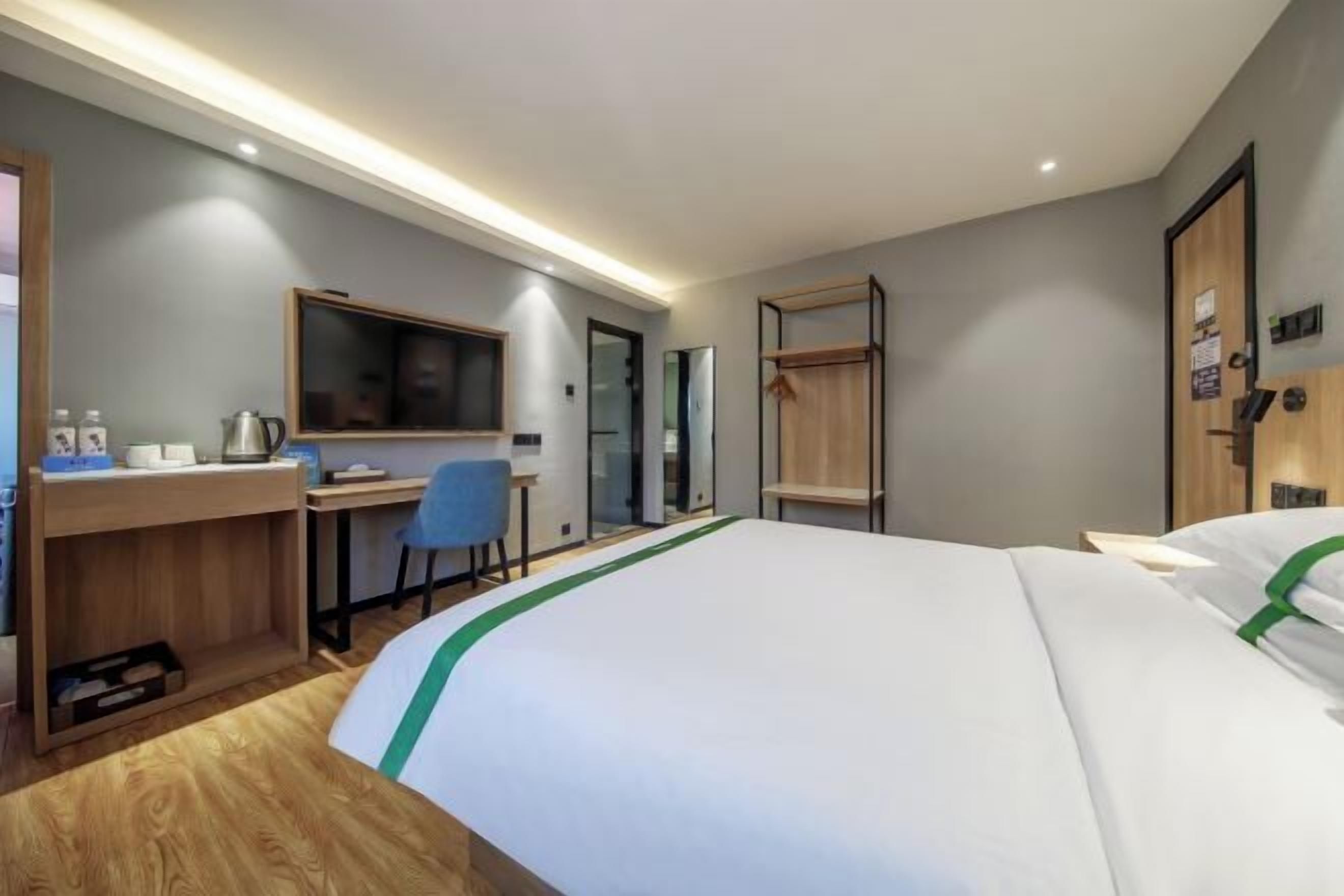 A modern hotel room with a large bed, a desk with a chair, a television, and a wardrobe.