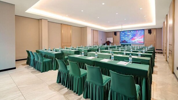 Un salón de conferencias con varias mesas y sillas, equipado con botellas de agua y documentos, y una pantalla grande al frente.