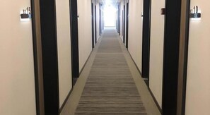 En hotellkorridor med dörrar numrerade från 10 till 15, ett golv täckt av matta och en rak gång som leder till en väl upplyst utgång.