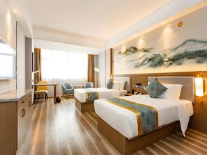 A hotel room with two beds, a desk, chairs, a large window, and a wall painting.