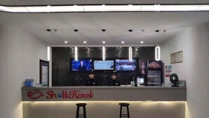 A hotel reception area with a modern design, featuring a reception desk, two barstools, and a flat-screen TV displaying news. - Shell Cangzhou Nanpi County Hospital of Traditional Chinese Medicine Hotel (Cangzhou)