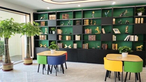 A modern library with green shelves, books, and decorative items. There are two tables with colorful chairs. - Shell Cangzhou Nanpi County Hospital of Traditional Chinese Medicine Hotel (Cangzhou)