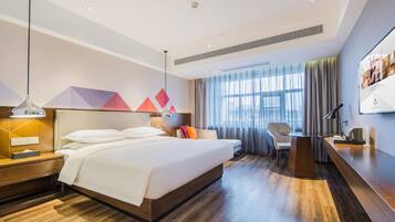 A modern hotel room with a large bed, a desk, a chair, and a view of the city through the window.