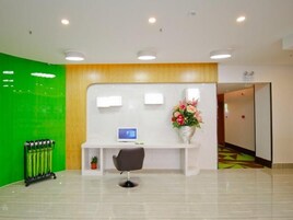 Zona de recepción con mostrador, una computadora, una silla, un florero con flores y una pared verde.