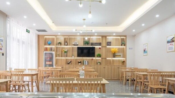 Un comedor con mesas y sillas de madera, una barra de buffet con recipientes de acero inoxidable, y un televisor montado en la pared.