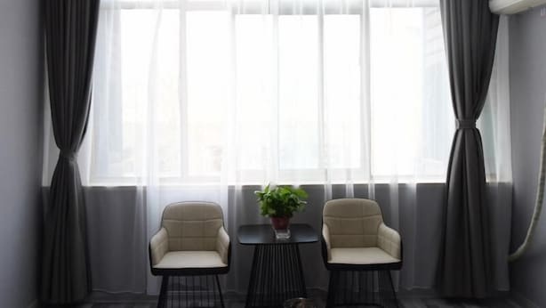 Een moderne kamer met twee stoelen en een klein tafeltje, grote ramen met dunne gordijnen, en een airconditioning op de muur.