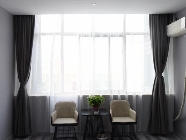 Une pièce moderne avec deux fauteuils et une petite table, de grandes fenêtres avec des rideaux légers et un climatiseur fixé au mur.