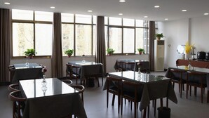 Een eetruimte met verschillende tafels en stoelen, grote ramen met gordijnen en potplanten.
