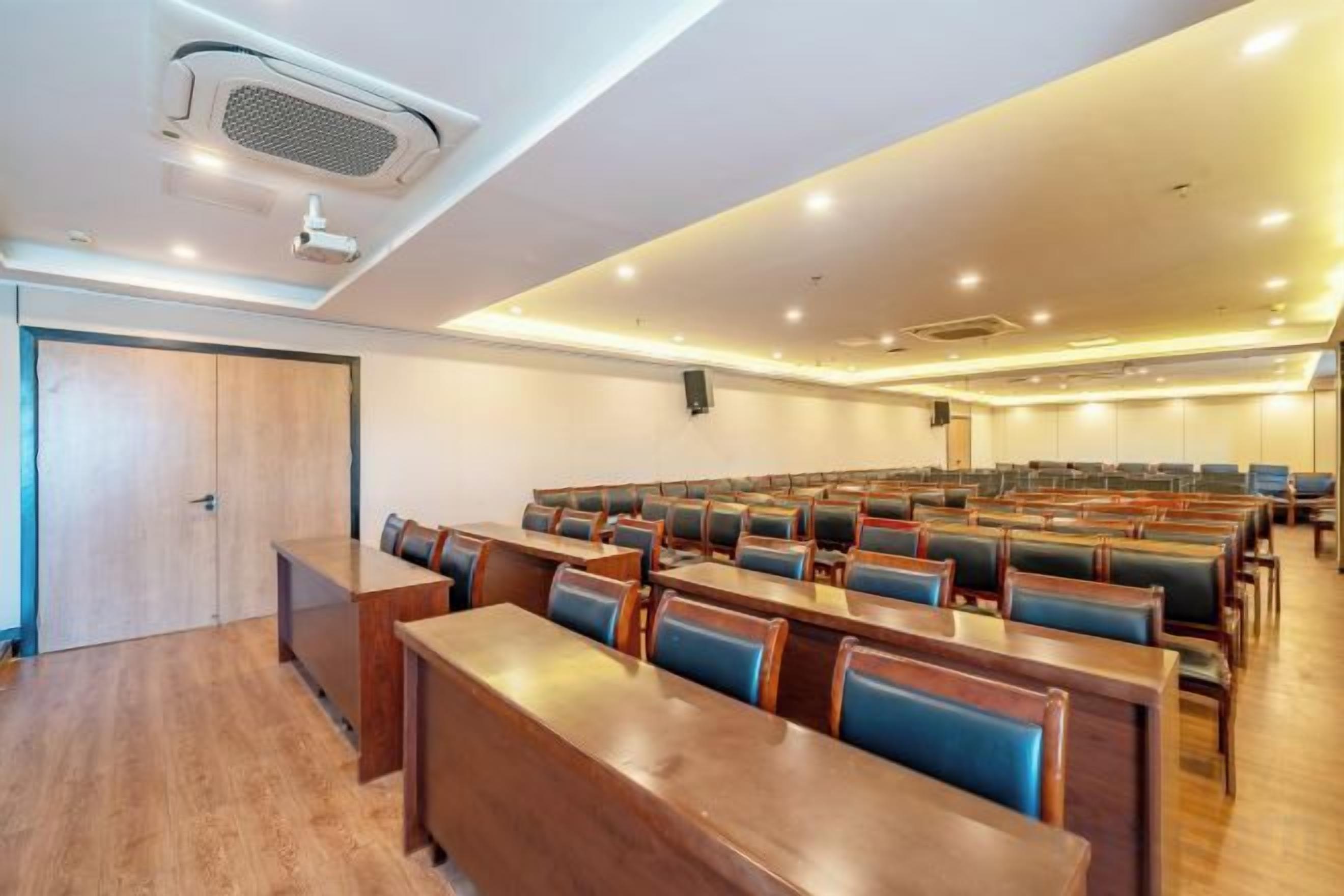 A conference room with rows of wooden tables and blue cushioned chairs, a projector, and a speaker on the wall.