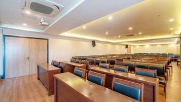 A conference room with rows of wooden tables and blue cushioned chairs, a projector, and a speaker on the wall.