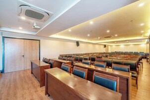 Un salón de conferencias con hileras de mesas de madera y sillas tapizadas en azul, un proyector y un altavoz en la pared.