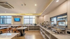 A modern dining area with wooden flooring, tables, and chairs. There are buffet stations with various food options. A television is mounted on the wall, and the room has large windows.