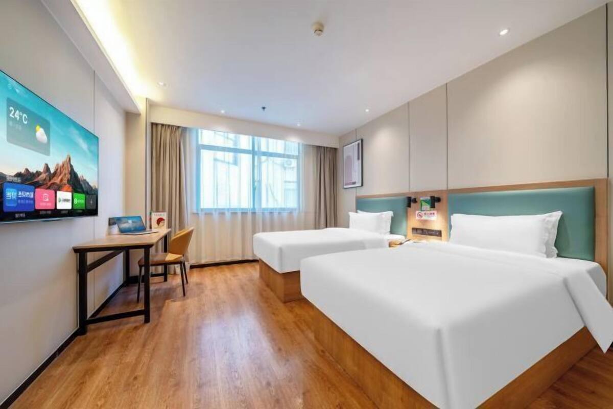 A hotel room with a wooden floor, two beds, a desk, a chair, a TV, and a window with curtains.