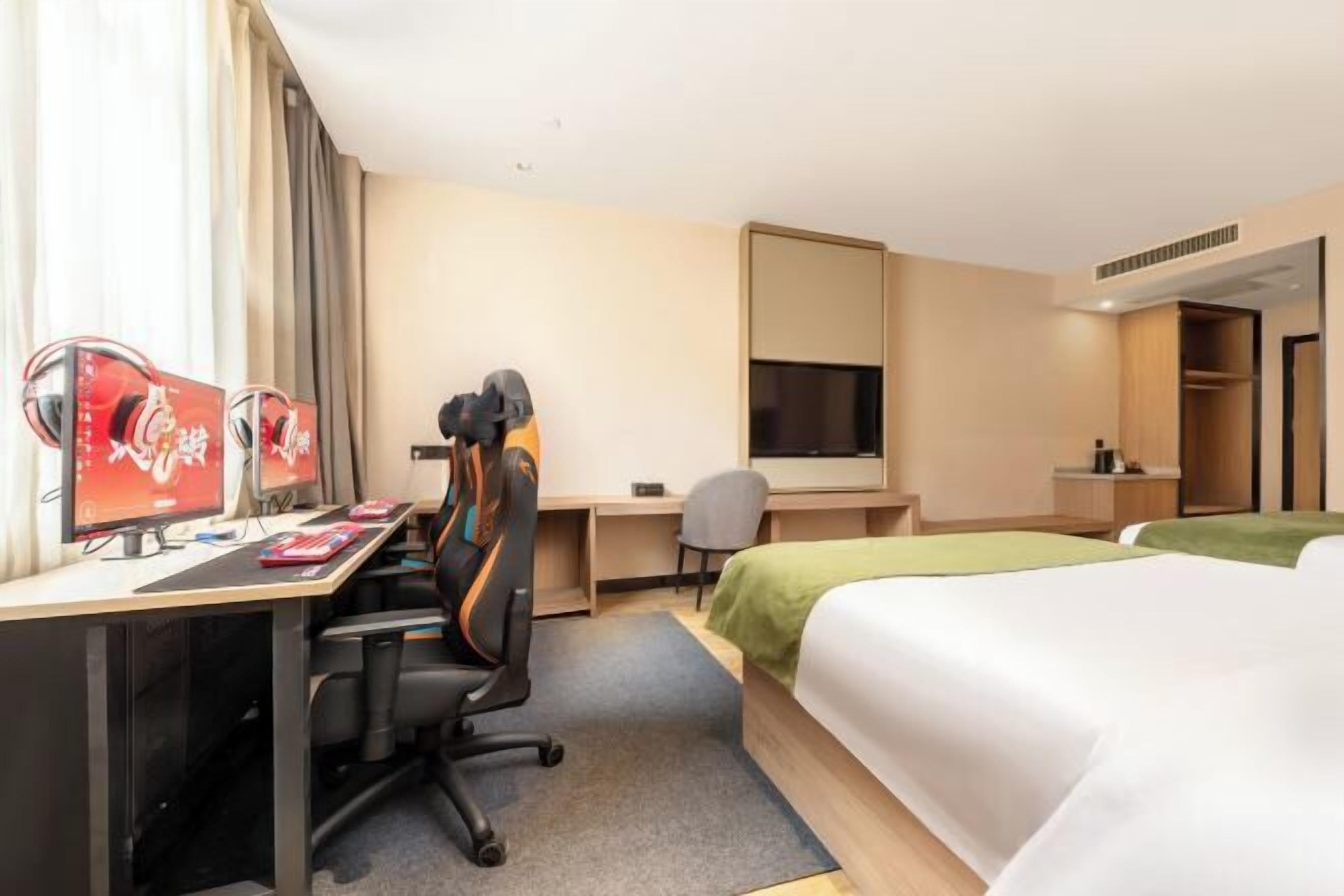 A hotel room with a desk, two monitors, a chair, a bed, and a wardrobe.