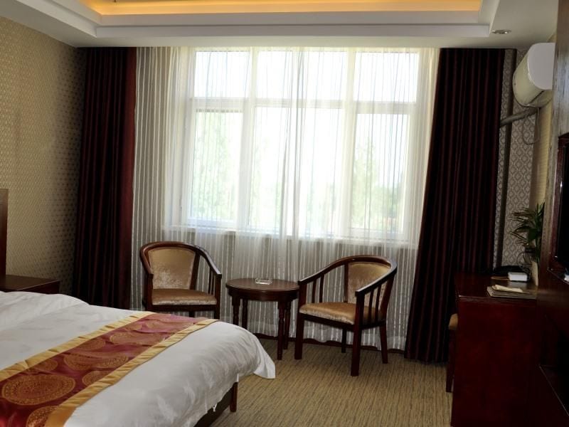 Een hotelkamer met een groot bed, twee stoelen, een tafeltje, een televisie en een raam met gordijnen.