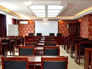 Un salón de conferencias con filas de sillas y mesas de madera, una pizarra y una pantalla de proyector.