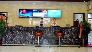 Área de recepção de hotel, com balcão de mármore, dois recepcionistas e uma grande tela de televisão exibindo conteúdo.