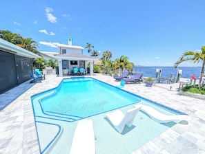 A swimming pool with lounge chairs and a covered seating area, surrounded by palm trees and a view of the sea.