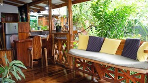 Une terrasse en bois avec un banc et des chaises, un coin cuisine équipé d’un réfrigérateur, et un jardin luxuriant et verdoyant que l’on aperçoit à travers le feuillage.