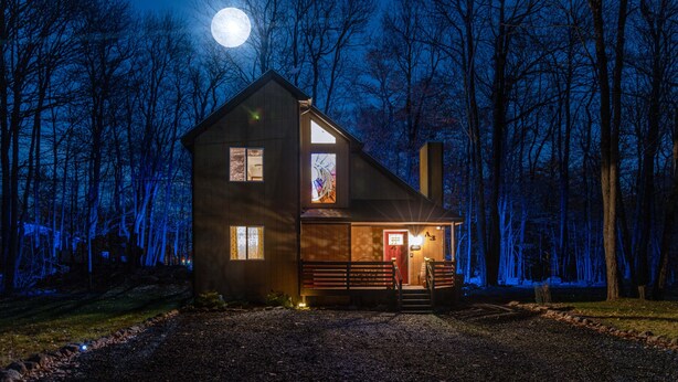 Ein zweistöckiges Holzhaus mit einer Veranda, einer roten Tür und einem beleuchteten Fenster.