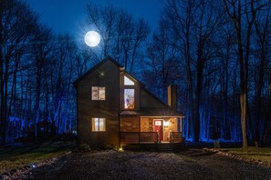 A two-story wooden house with a front porch, a red door, and a lit window. - The Dragon House, Hot Tub, Mini Golf, Pet Friendly (Tobyhanna)