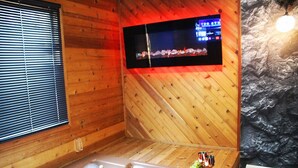 Ein Holzhäuschen im Inneren mit Holzboden, einem großen Fenster mit Jalousien, einem an der Wand befestigten Fernseher und einer Steinwand.