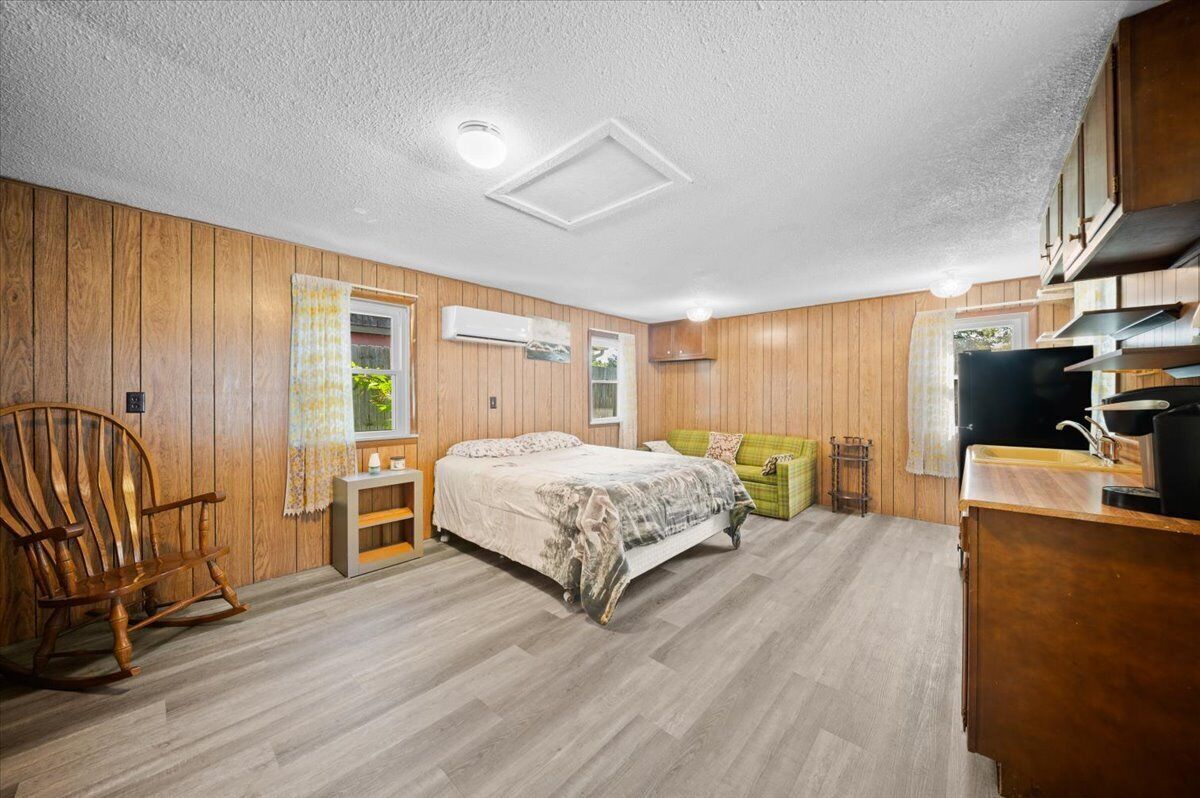 A room with wooden paneling, a bed with a patterned bedspread, a rocking chair, a green sofa, a small table, and a kitchenette.