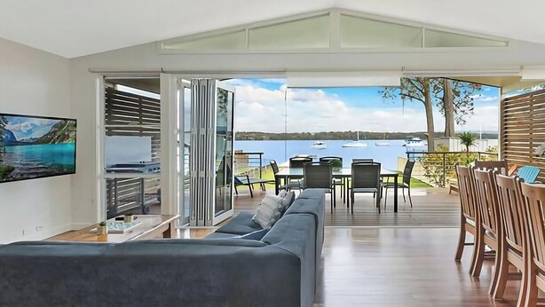 A modern living room with a large sofa, dining table, and chairs, a TV, and a view of the sea. - Sunset Central Coast - Lake, Kayaks & Jetty! (Mannering Park)