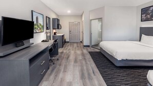 A modern hotel room with a large flat-screen TV, a bed with white bedding, a desk with a chair, and a small kitchenette.