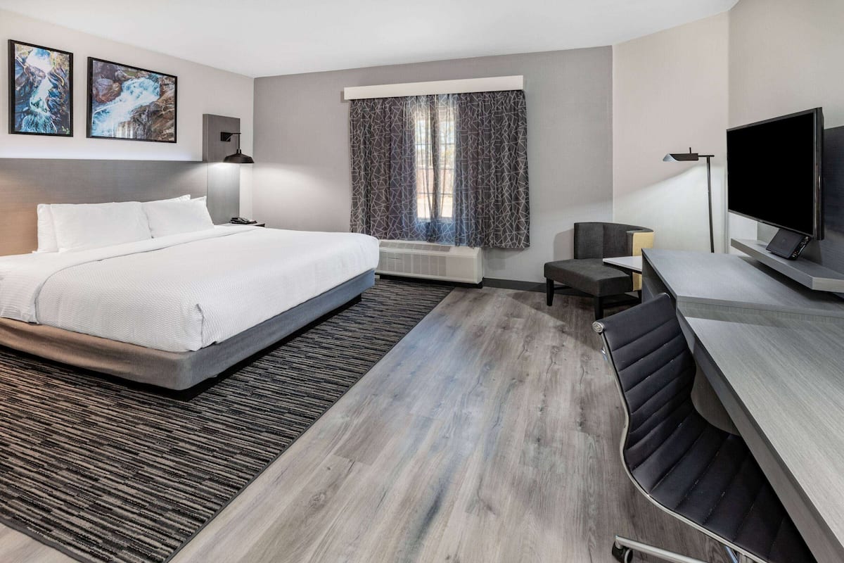 A modern hotel room with a large bed, a desk with a flat-screen TV, a small table, and a chair.