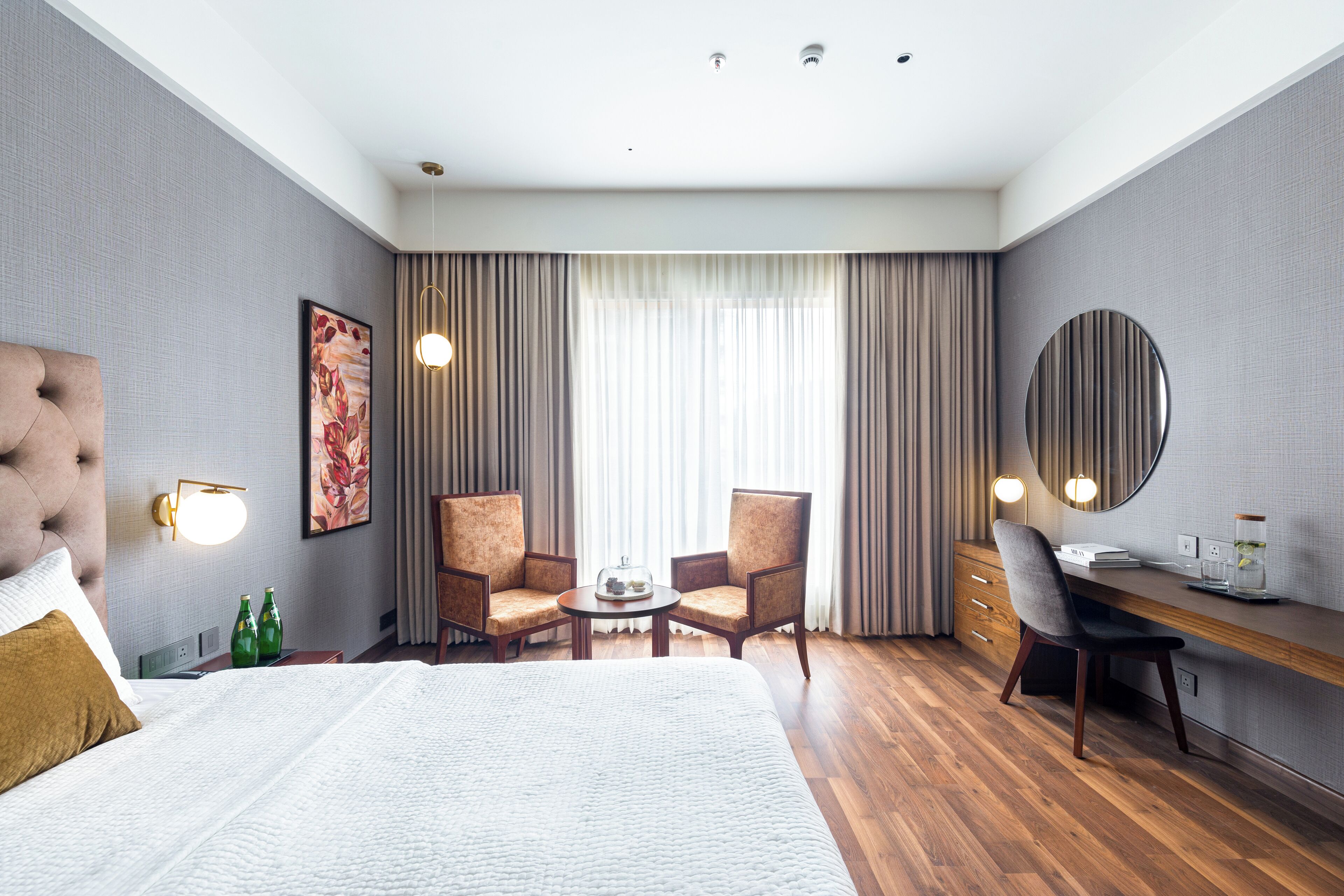 A hotel room with a large bed, two armchairs, a desk, and a round mirror.