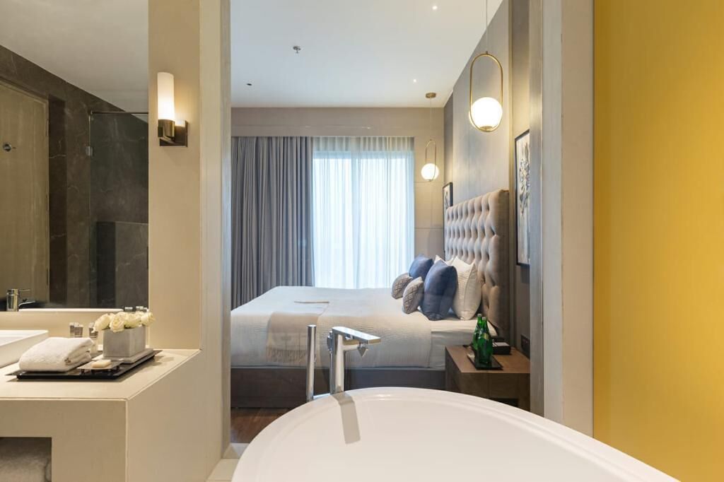 Ein modernes Hotelzimmer mit einem großen Bett, einem separaten Badezimmer inklusive Badewanne, einem Schminktisch mit Spiegel und einem großen Fenster mit Vorhängen.