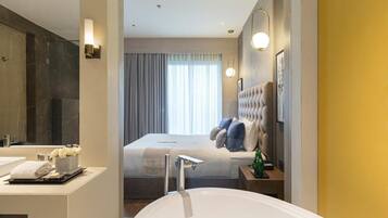 A modern hotel room with a large bed, a separate bathroom with a bathtub, a vanity with a mirror, and a large window with curtains.