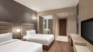 A modern hotel room with two beds, a TV, and a bathroom with a glass shower enclosure.
