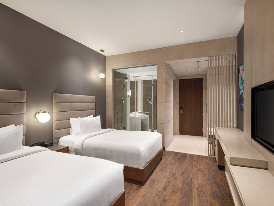 A modern hotel room with two beds, a TV, and a bathroom with a glass shower enclosure.
