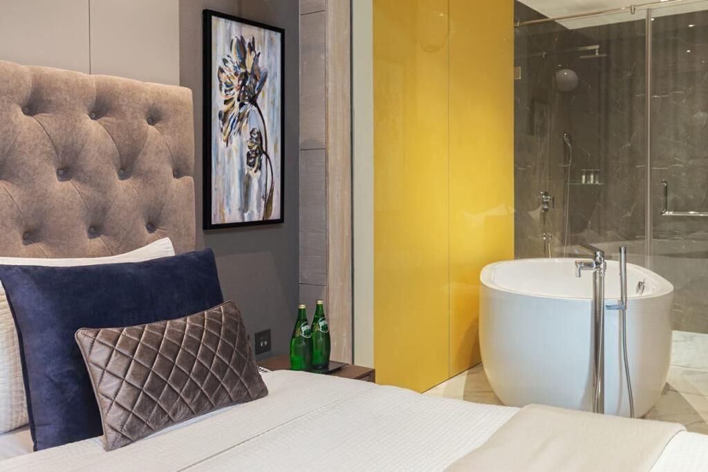 Ein modernes Schlafzimmer mit einer gesteppten Kopfteil, einem großen Bett mit Kissen, einem gerahmten Kunstwerk, einer gelben Akzentwand, einer Duschkabine aus Glas und einer weißen Badewanne.