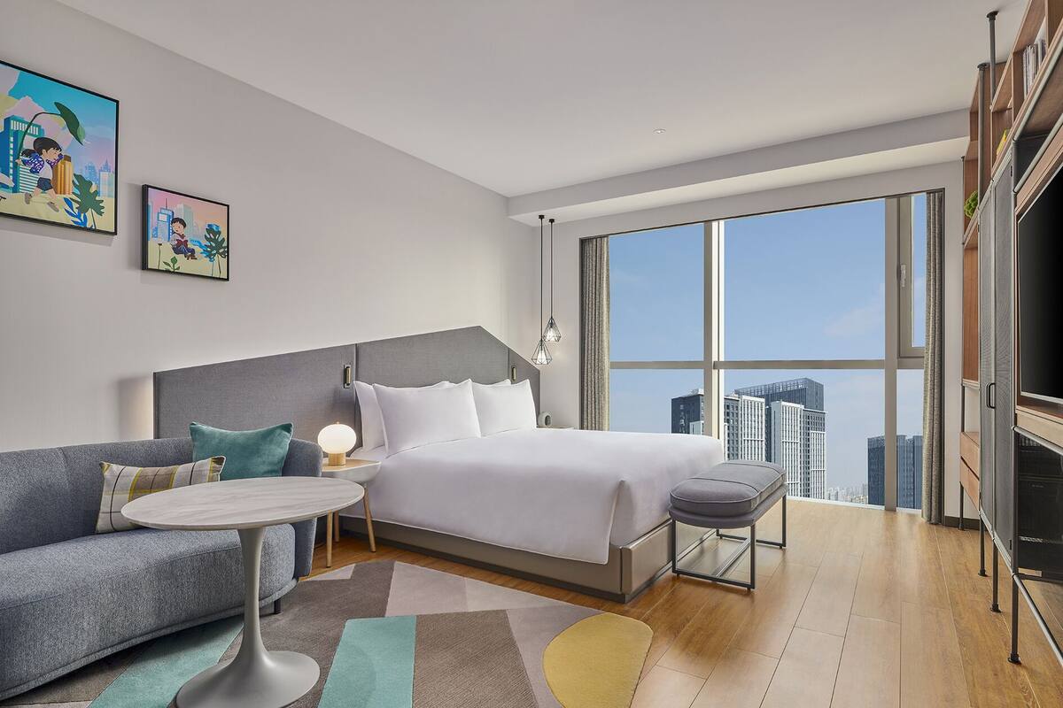 Une chambre d’hôtel moderne dotée d’un grand lit, d’un canapé, d’une petite table et offrant une vue sur la ville grâce à de grandes fenêtres.