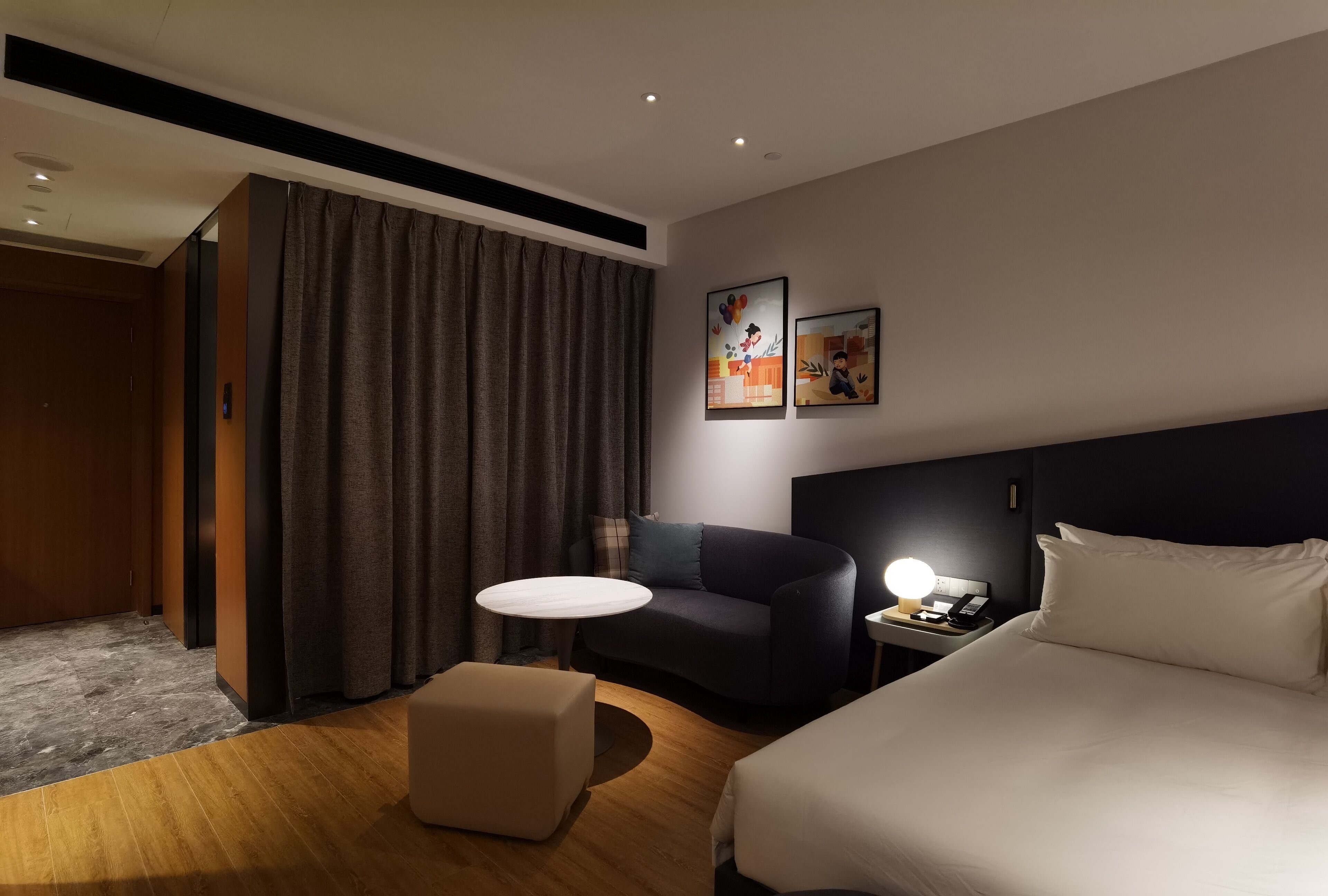 A hotel room with a bed, a desk with a lamp, a chair, a small table, and a large window with curtains.