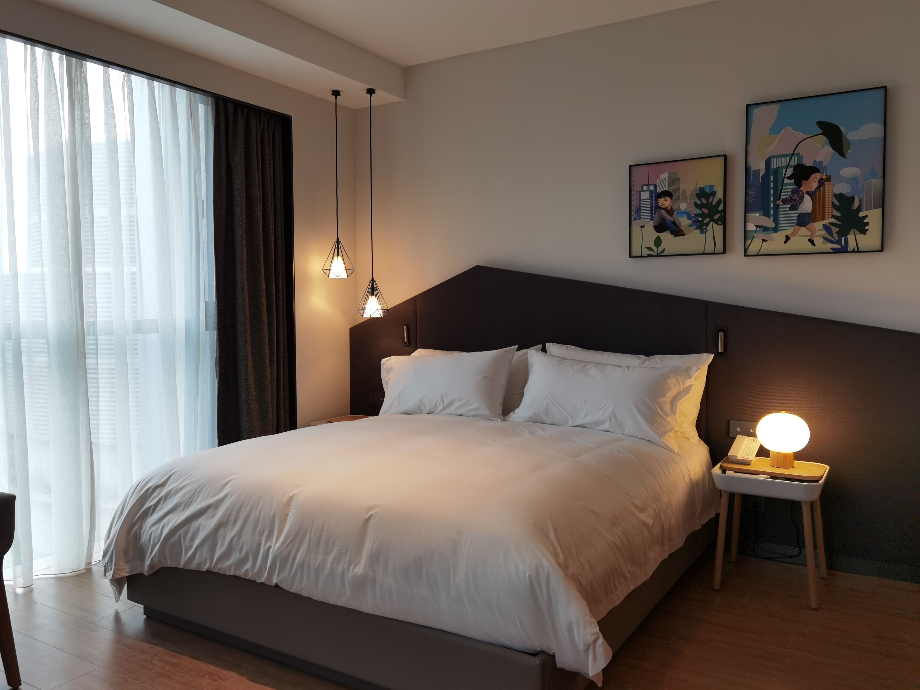 A modern hotel room with a large bed, two pendant lights, a bedside table with a lamp, and artwork on the wall.