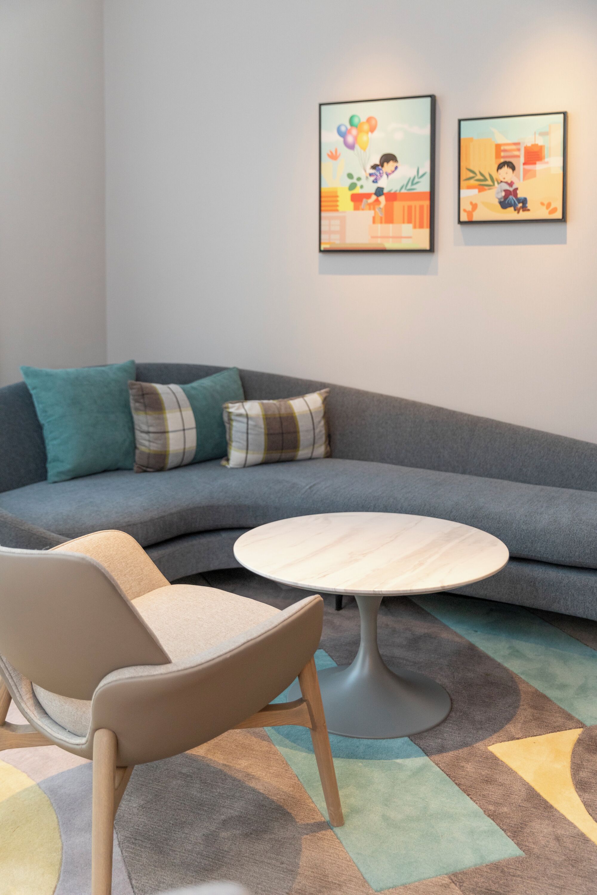 Un salon moderne avec un canapé d’angle gris, une table basse ronde et un tapis à motifs. Deux œuvres d’art encadrées ornent le mur.