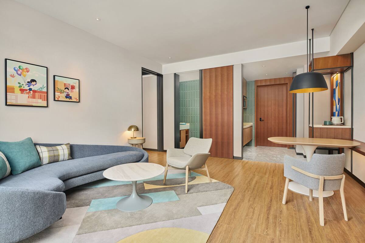 Un salon moderne avec un canapé d’angle, une table basse ronde et un coin repas comprenant une table et des chaises. Deux œuvres d’art encadrées ornent le mur, ainsi qu’une porte en bois et une vue sur une autre pièce.